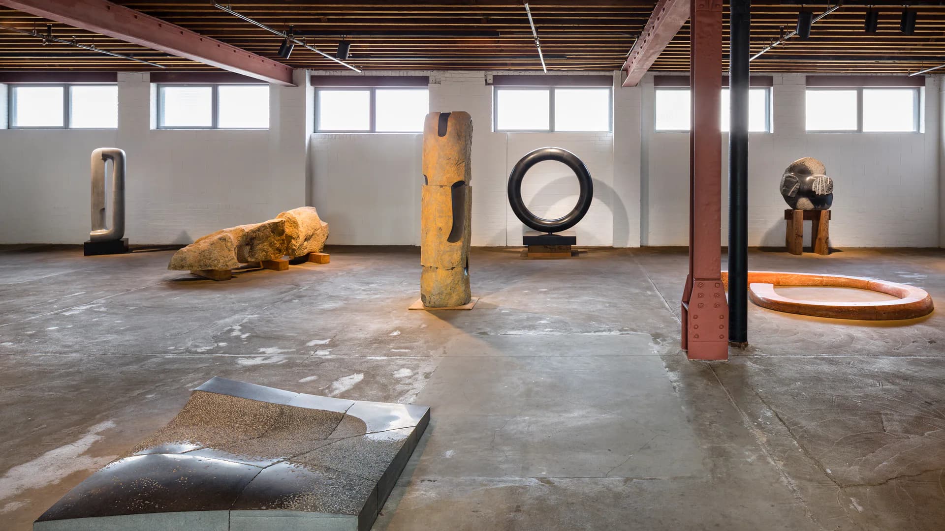 Industrial gallery space with concrete floor displaying abstract sculptures including wooden forms, a tire, and cylindrical shapes arranged throughout under skylights and exposed beams.