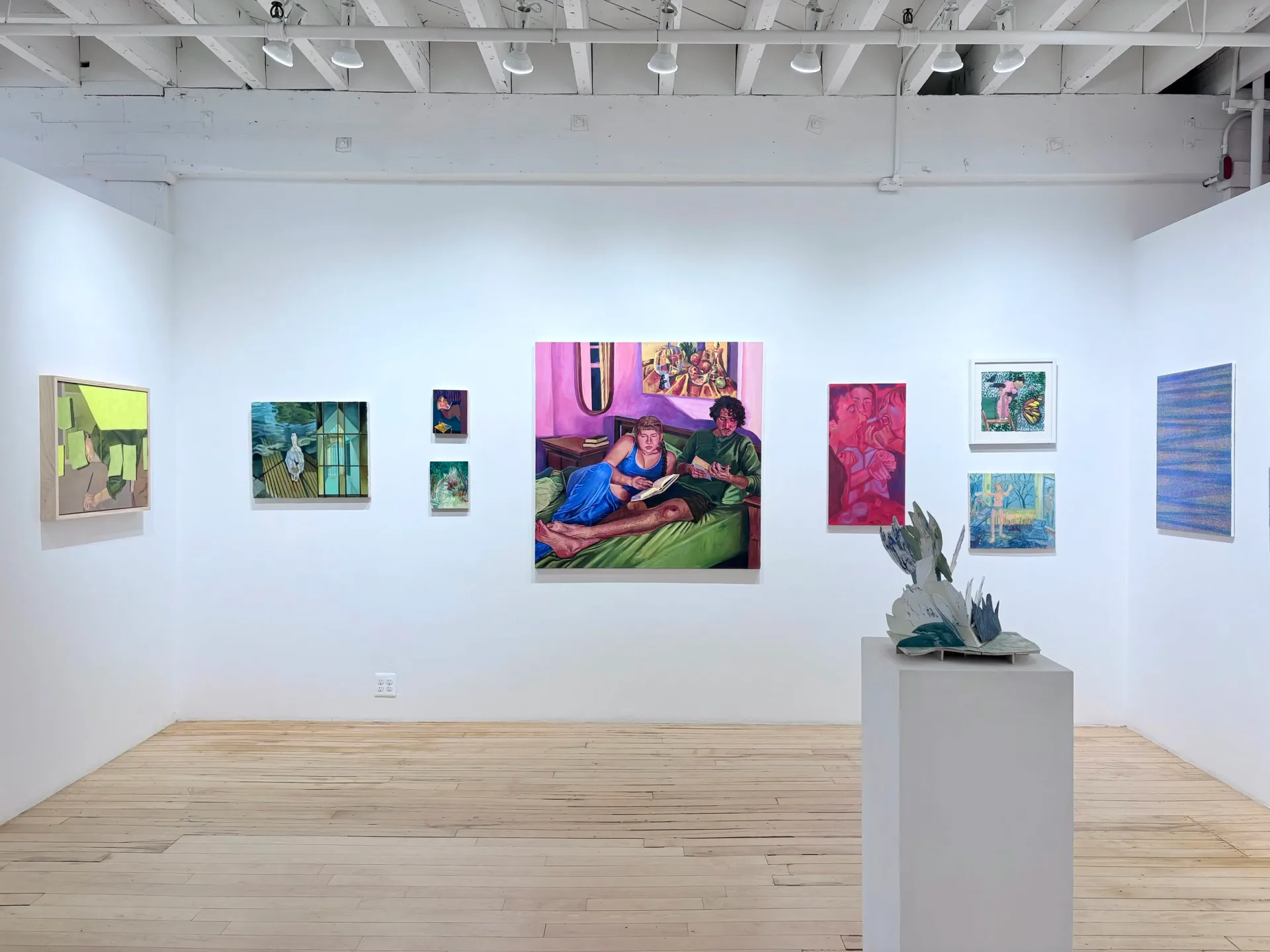 The Painting Center gallery interior with light wood floors, white walls, and various colorful paintings hung at varying heights; a white pedestal with potted plant stands in the center.