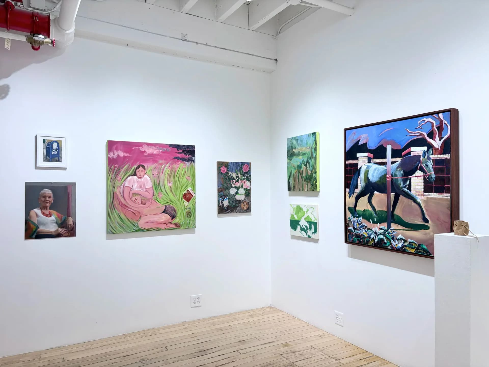 The Painting Center gallery corner with light wood floors, exposed white ceiling beams, and colorful artworks hung on white walls; a white pedestal with object stands in foreground.
