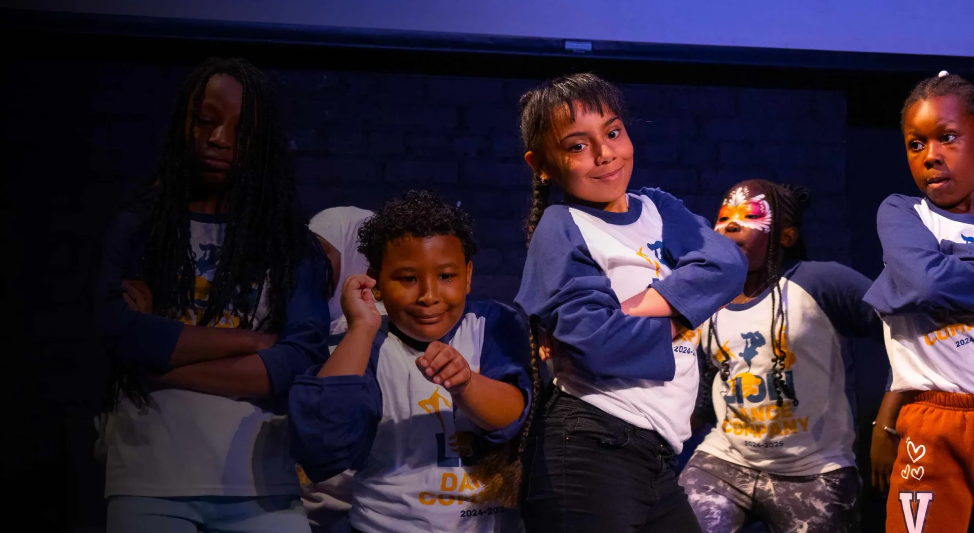 Children on stage wearing blue and white clothing, smiling and posing under blue stage lighting during what appears to be a performance or presentation.