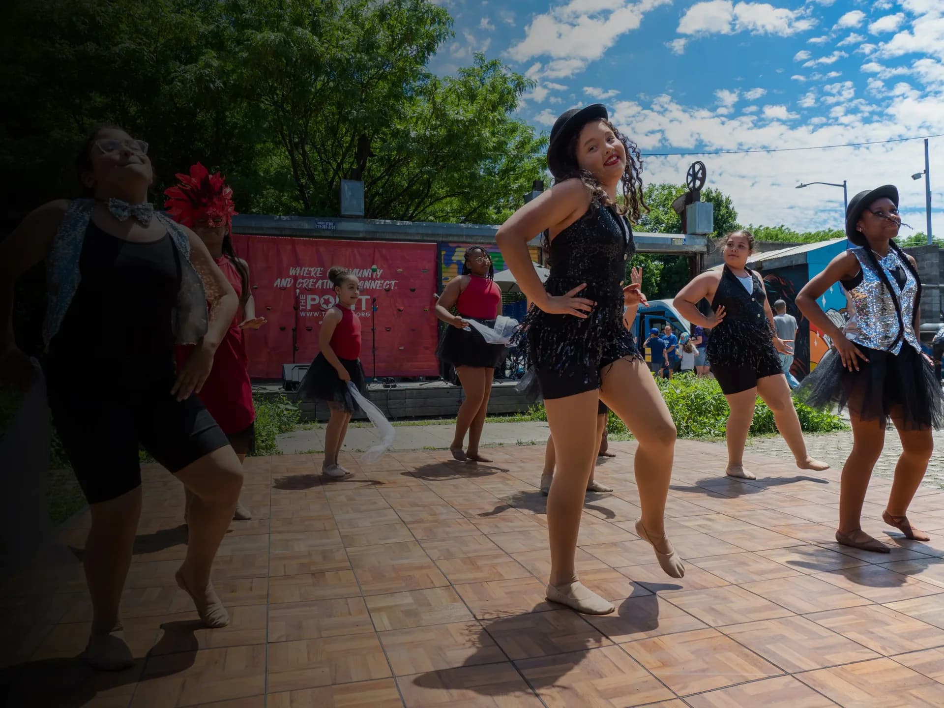 Children dancing outdoors on a paved surface with a red structure visible in the background, captured in bright daylight.