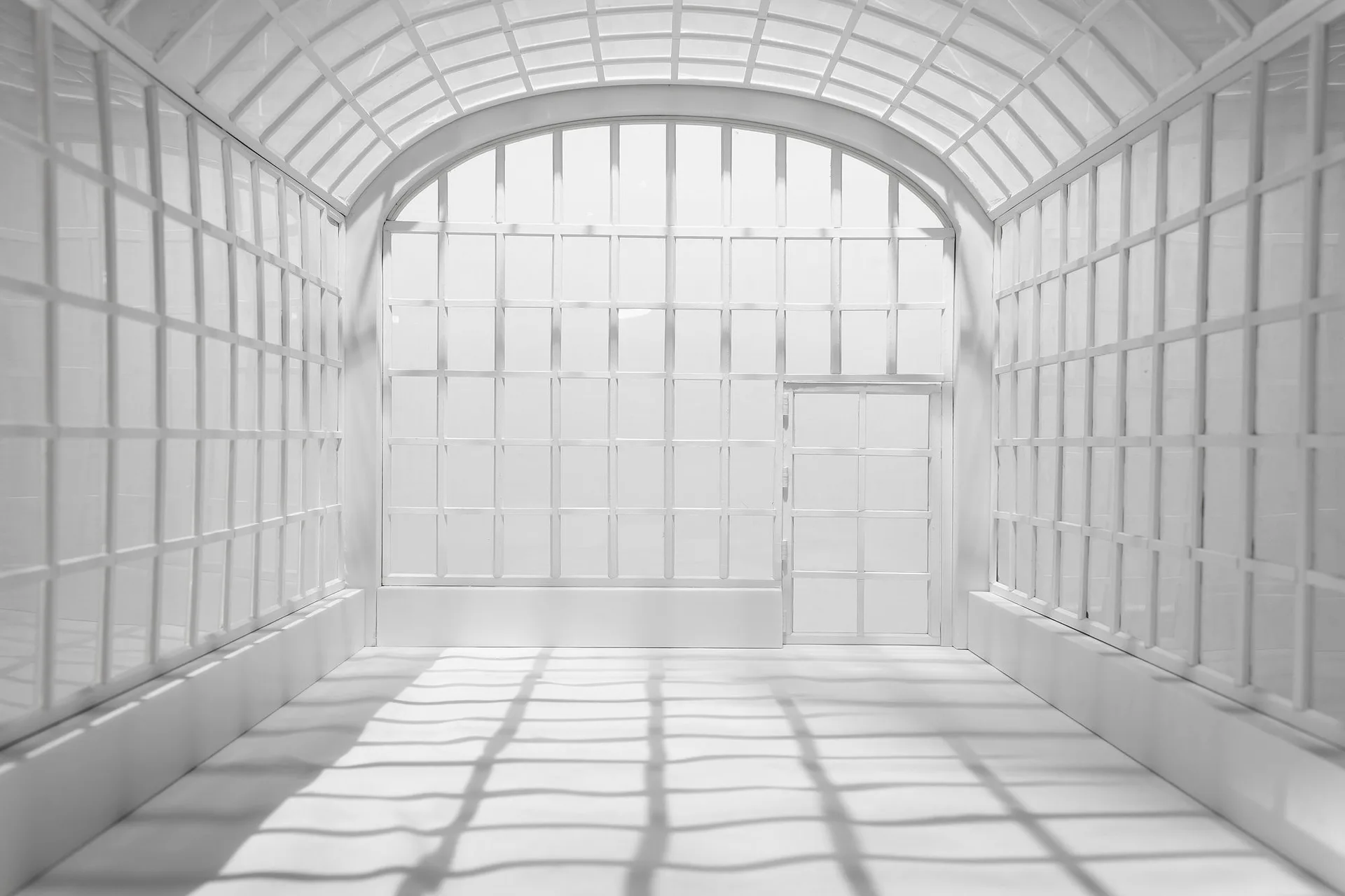 Bright, minimalist interior of an empty white gallery or studio space with gridded walls, barrel-vaulted ceiling, and natural light creating linear shadows on the floor.