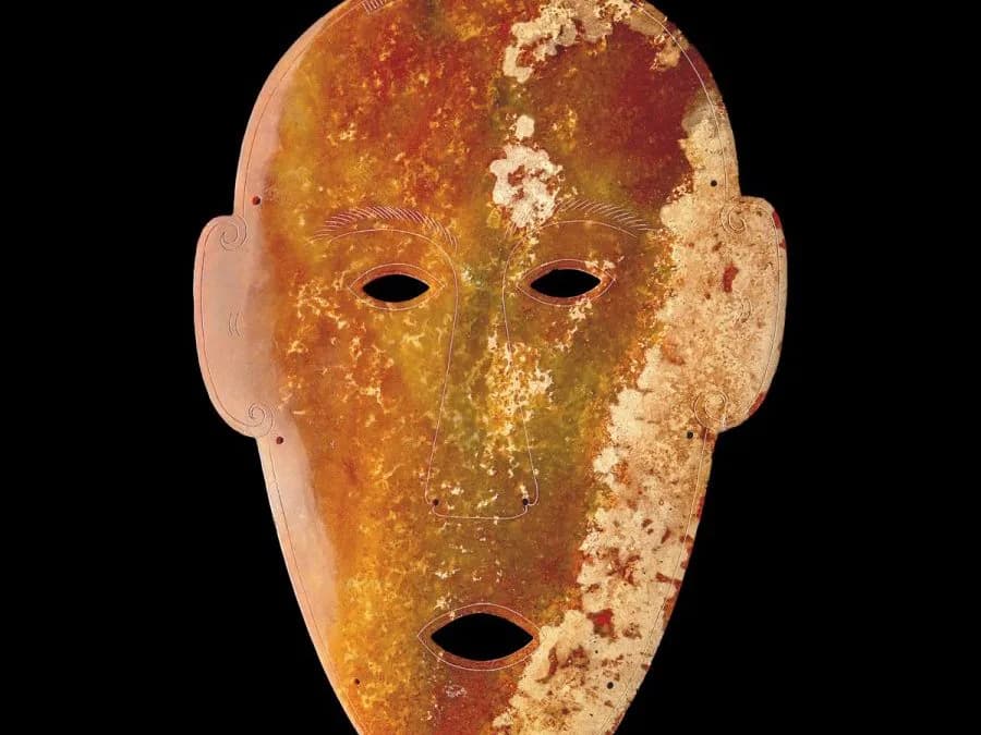 Ancient terracotta mask with geometric facial features, two eye holes and one mouth opening, oxidized orange and gold patina against black background.