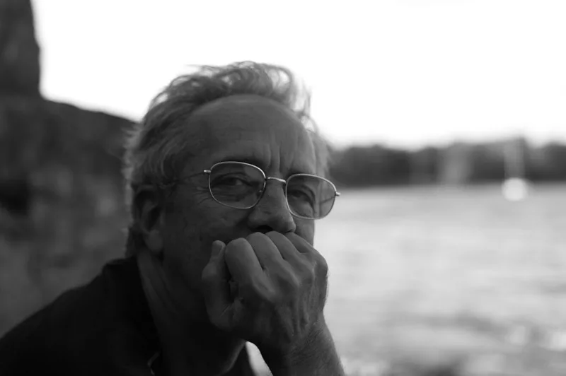 Black and white portrait of an older man wearing glasses and casual clothing, hand near face, contemplative expression beside a waterfront.