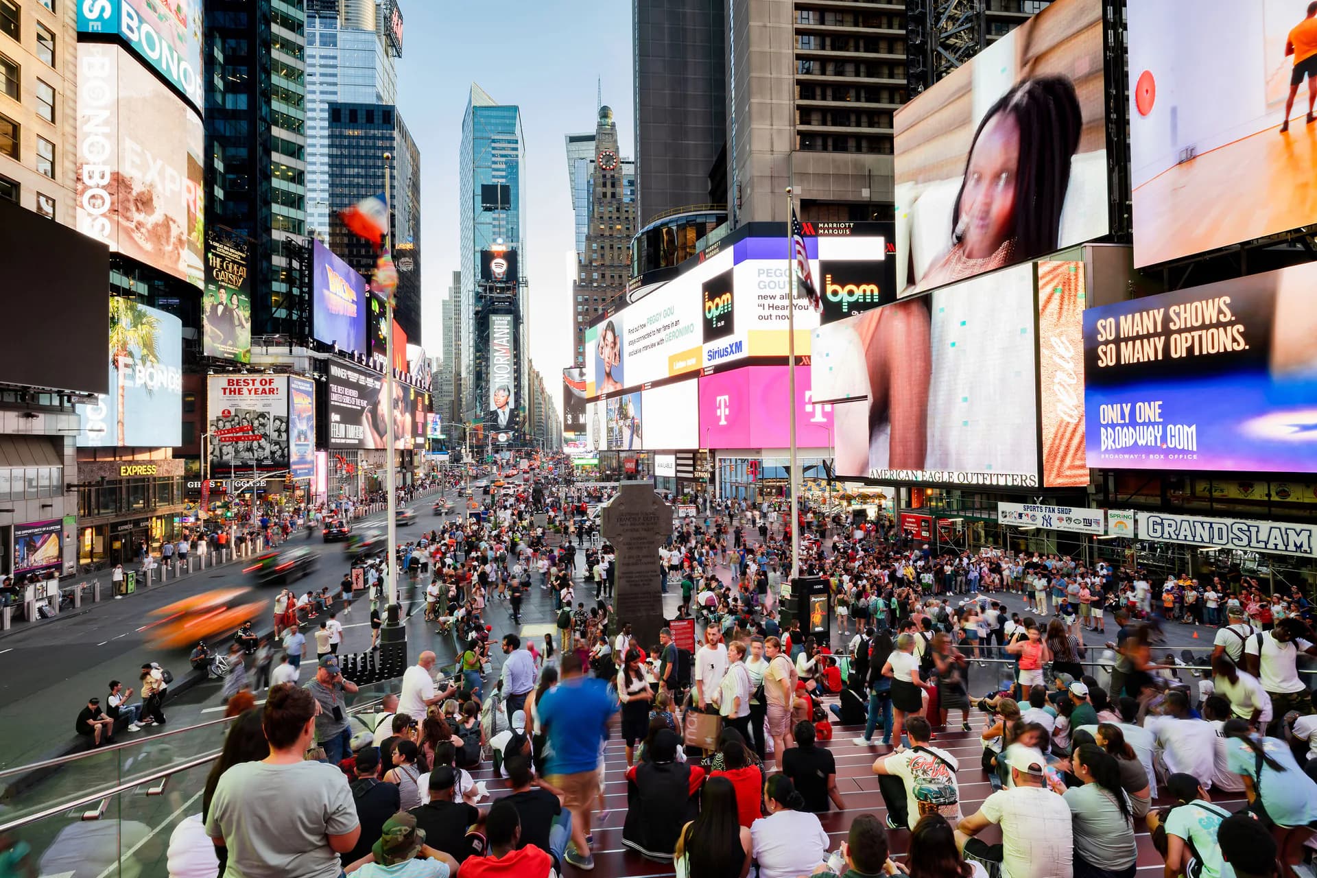 Crowded pedestrian plaza in Times Square with tall buildings, digital billboards, and masses of tourists on a clear day.