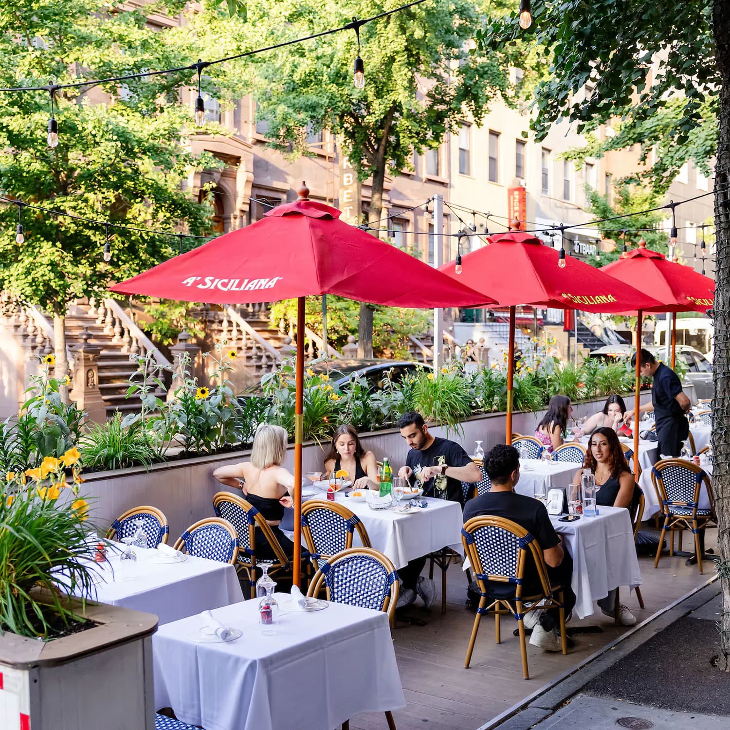 Outdoor dining terrace with white tablecloths, diners under red umbrellas, surrounded by potted plants and brick historic buildings.