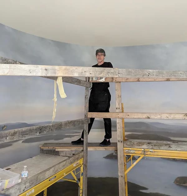 Person in black clothing standing on wooden scaffolding against a large-scale landscape mural in Timothy Brewer Studio.