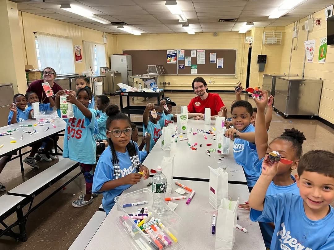 Classroom workshop at Toy Museum of New York with children in blue shirts working at white tables on craft projects, instructor in red shirt supervising.