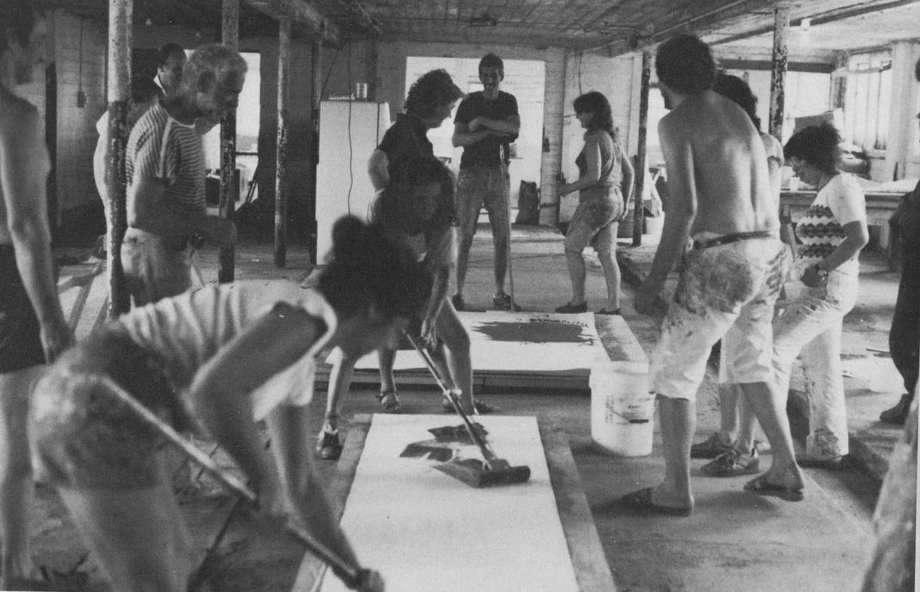 Black and white photograph of multiple people in what appears to be a studio or workshop space, gathered around artworks or materials on the floor.