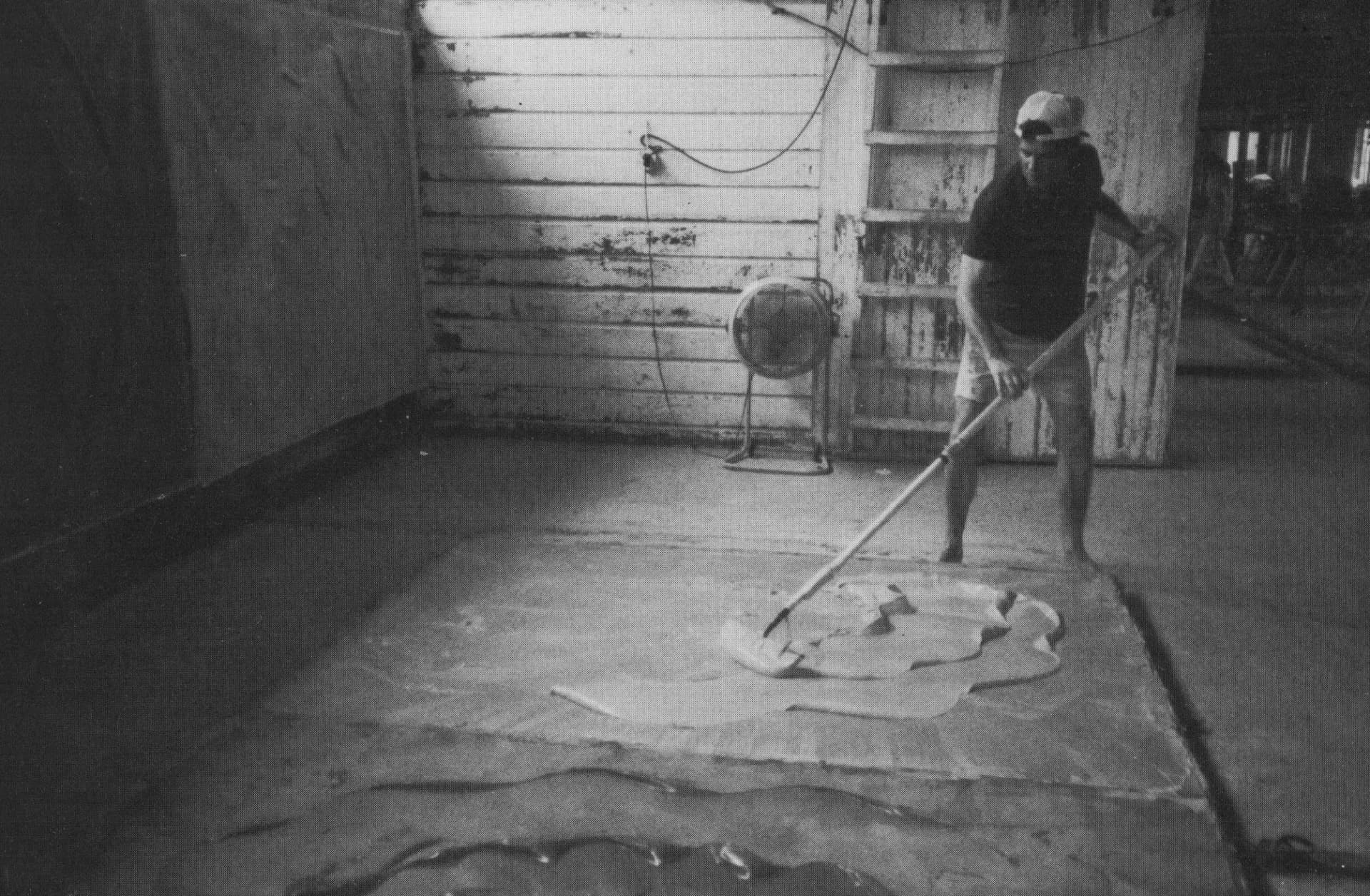 Black and white photograph of a person in an industrial or studio space with exposed walls and wiring, appearing to work on or view a piece on the ground.