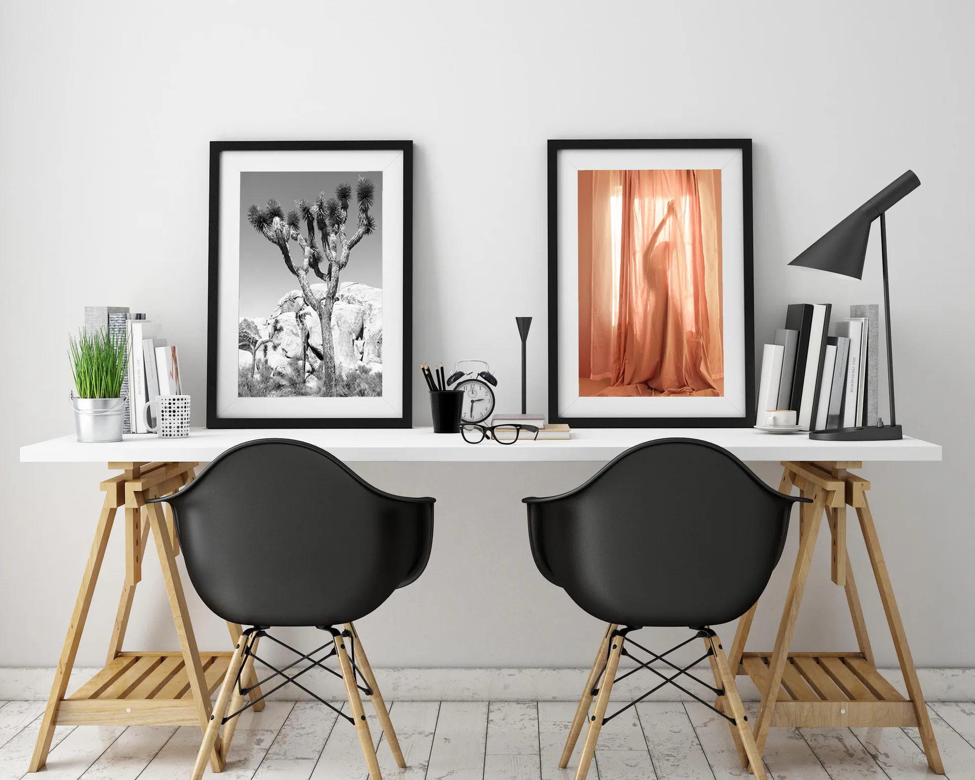 Workspace with white shelf holding two framed black-and-white artworks, books, lamp, and plants above wooden desk with black chairs.