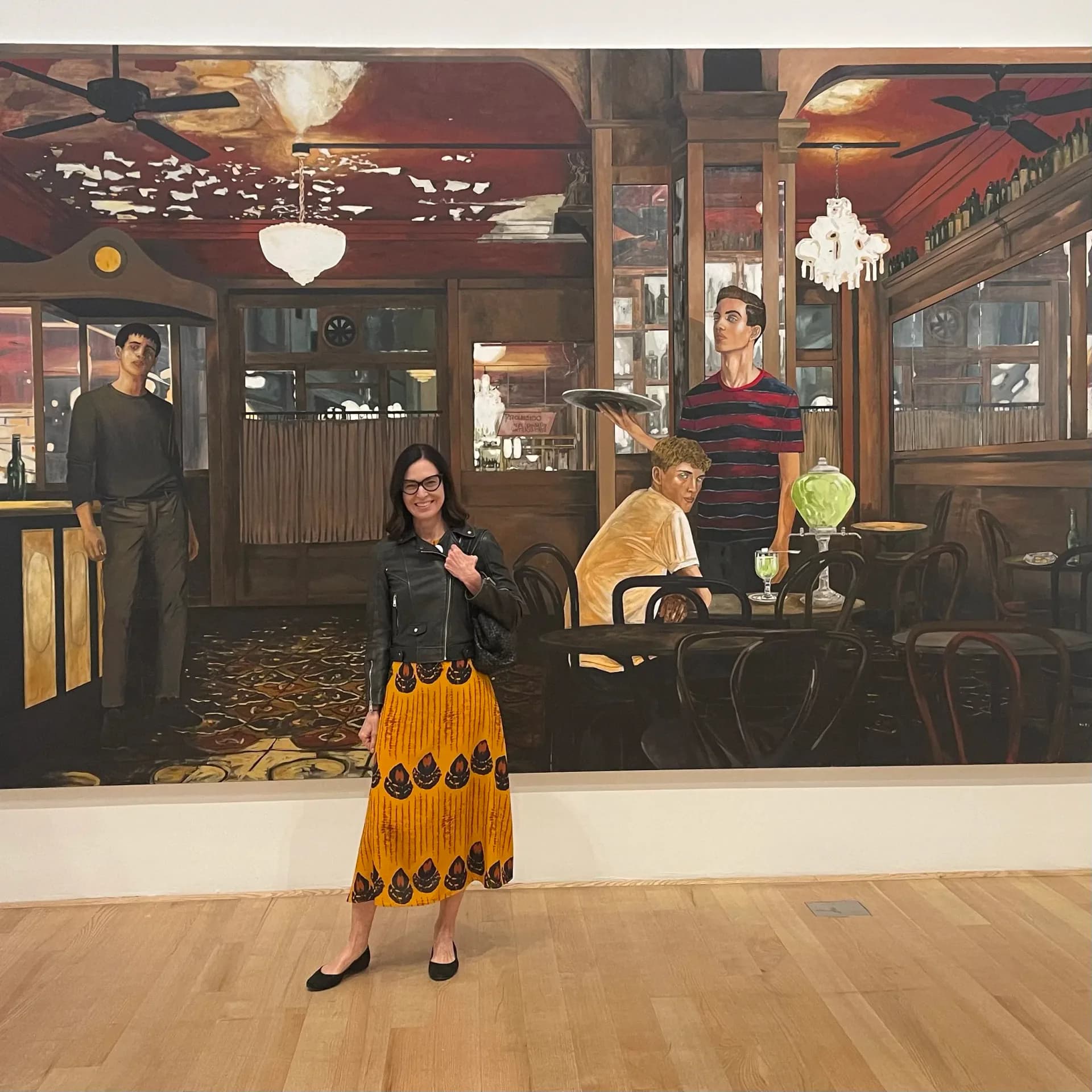 Woman in yellow skirt standing before large interior photograph of a vintage café or restaurant with warm lighting and wooden details.