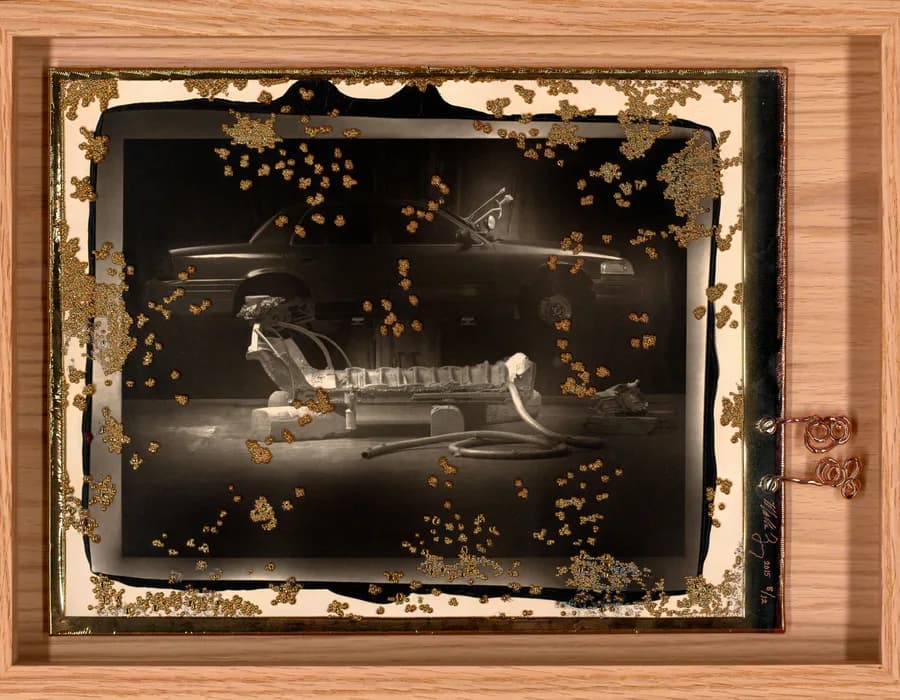 Mixed-media installation with distressed wood frame and white border surrounding dark interior with sculptural form and scattered gold spheres.