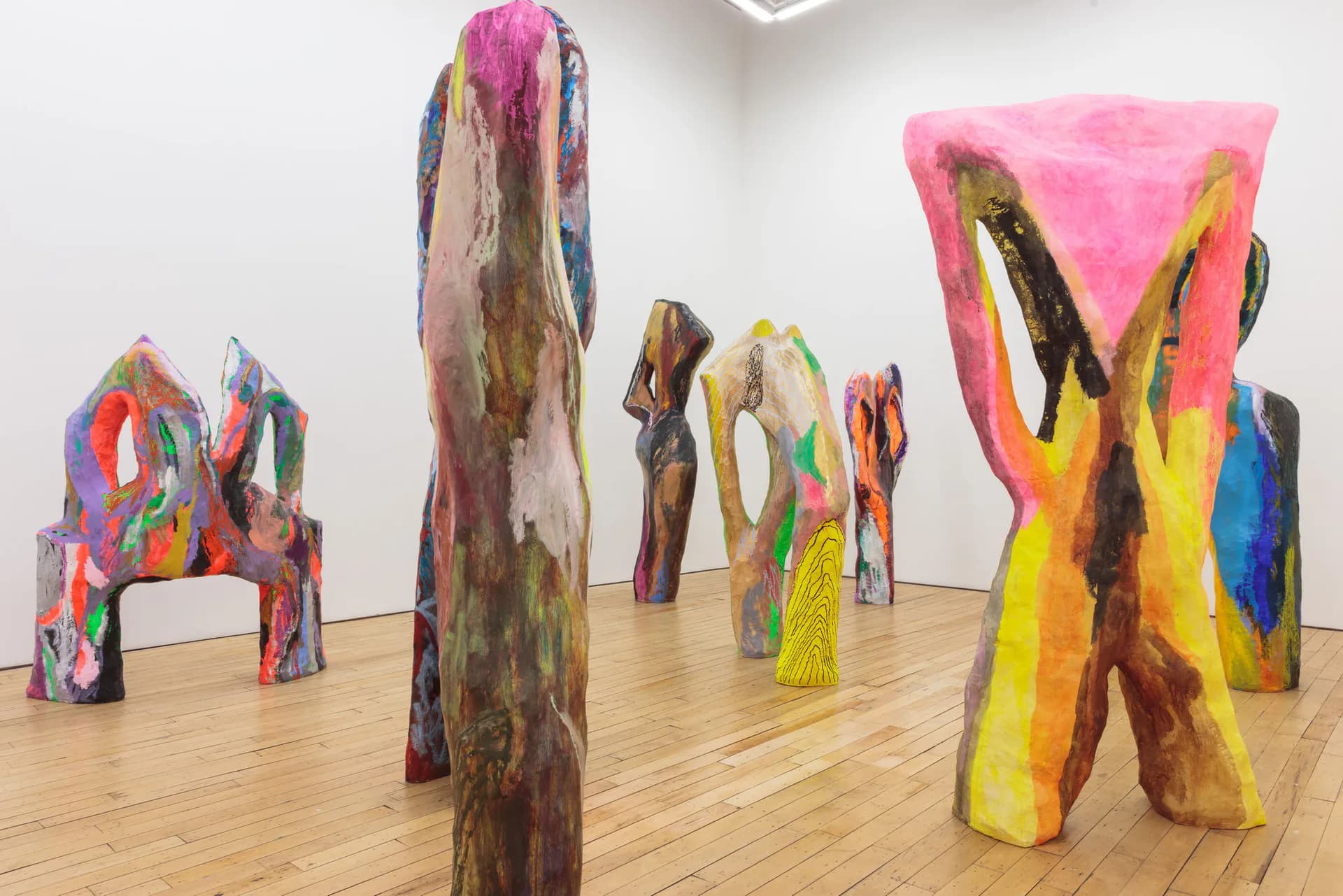 Gallery interior with wooden floor displaying multiple large, colorful sculptural forms in pink, yellow, blue, and natural wood tones arranged spatially.