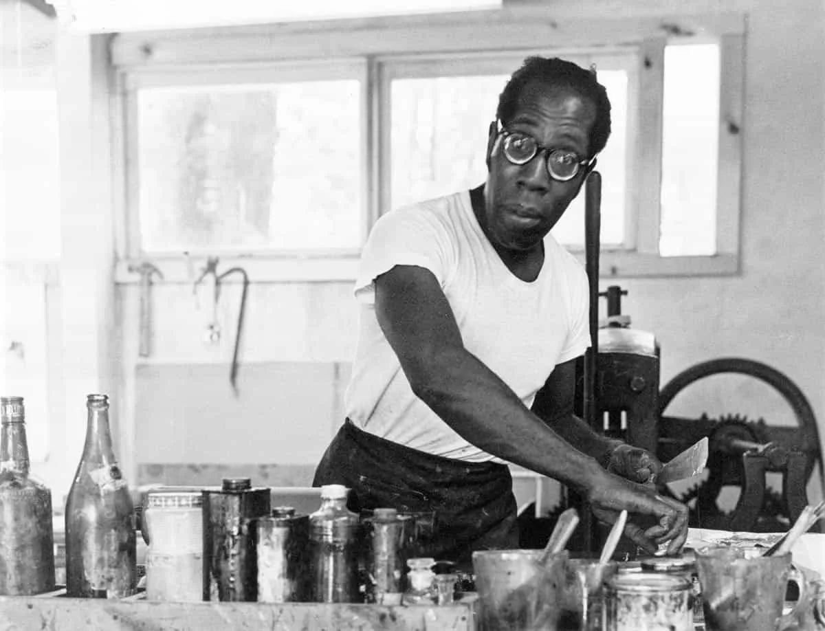 Black and white photograph of a man wearing glasses and light-colored shirt working at a table with jars and materials in a studio setting.