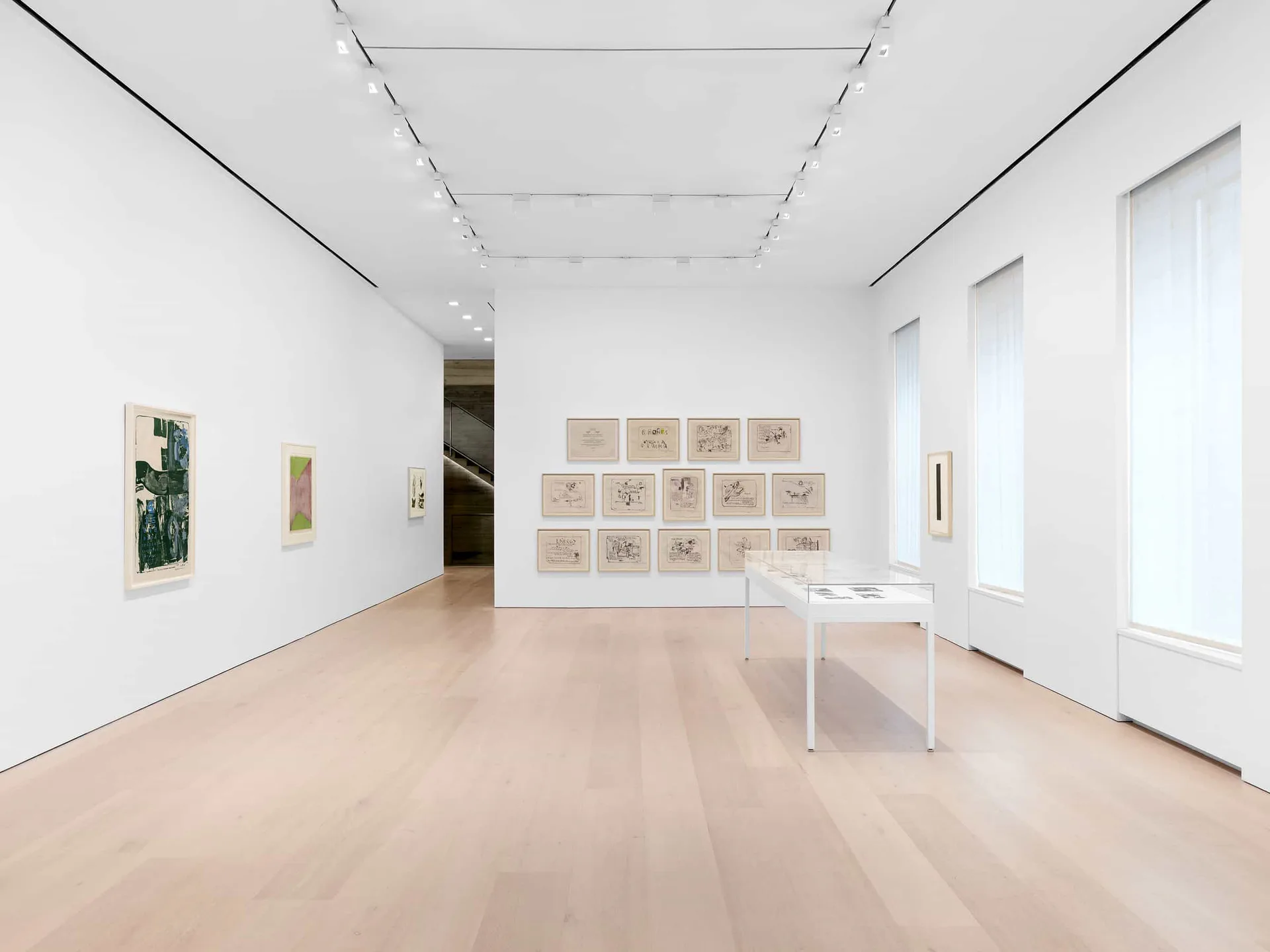 Minimalist gallery interior with white walls, light wood flooring, grid of framed works on right wall, small table, and natural light from windows.