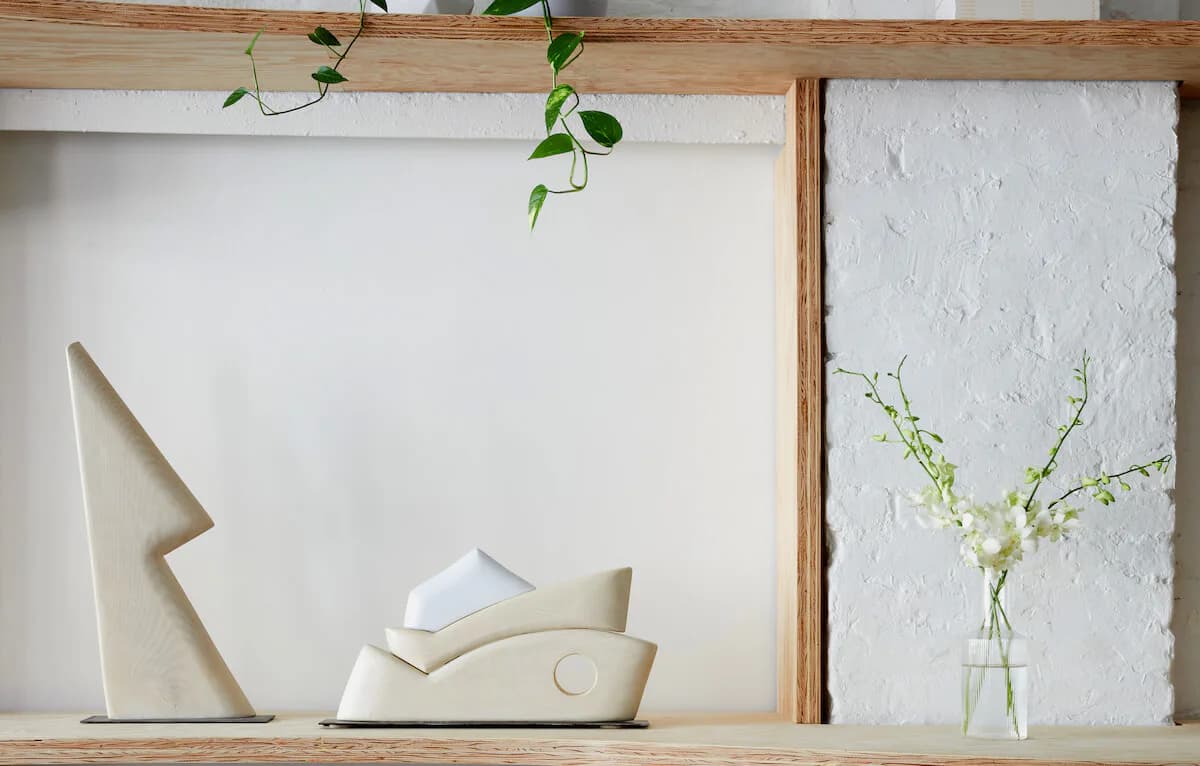 Gallery shelf display with white ceramic sculptural objects, a clear glass vase with green stems, wooden frame, and draped beige fabric against white walls.