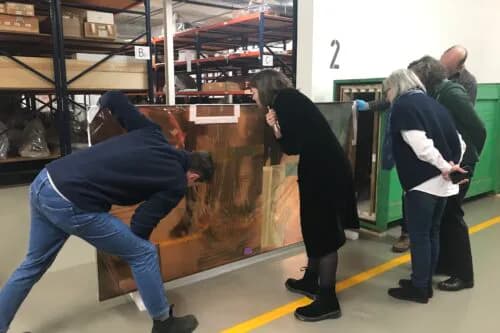 Gallery visitors viewing large artwork or installation on floor in industrial-style interior space with yellow line marking.