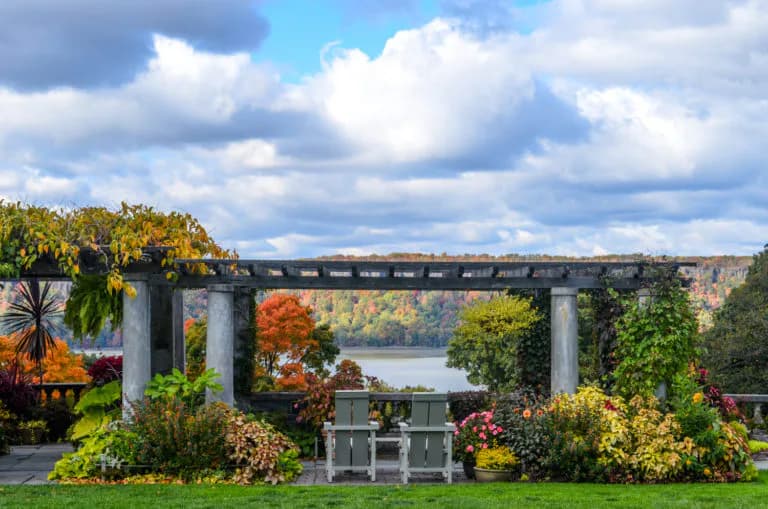 Stone pergola with Adirondack chairs overlooking autumn gardens and distant water at Wave Hill, colorful flowering shrubs in foreground beneath cloudy sky.