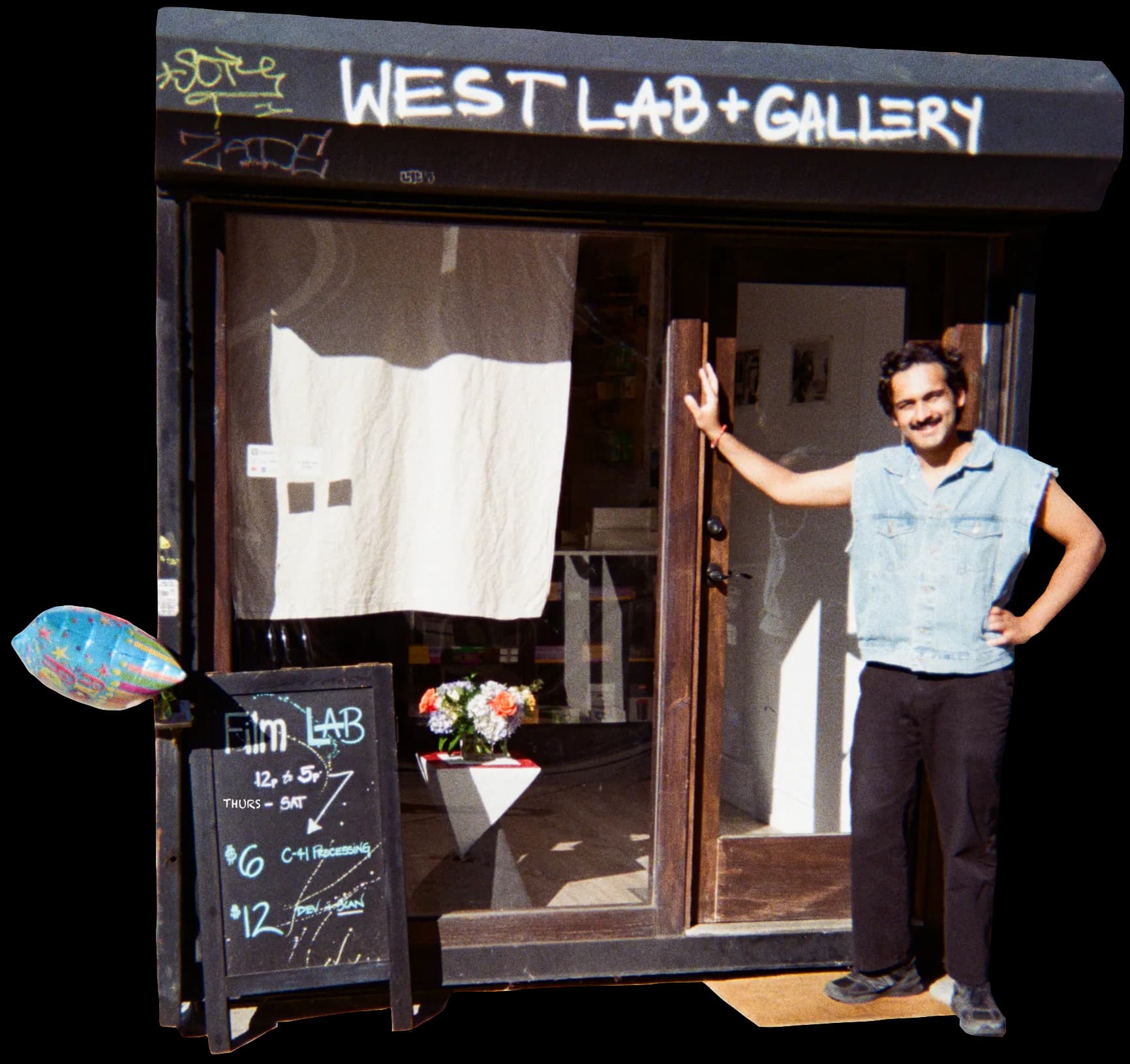 Daytime exterior photograph of Westlab + Gallery storefront with person standing in doorway, white artwork visible in window, flowers and small chalkboard sign at entrance.