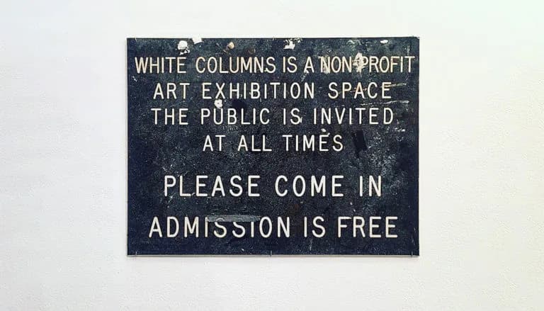 Dark blue sign with white text reading 'WHITE COLUMNS IS A NON-PROFIT ART EXHIBITION SPACE THE PUBLIC IS INVITED AT ALL TIMES PLEASE COME IN ADMISSION IS FREE'.