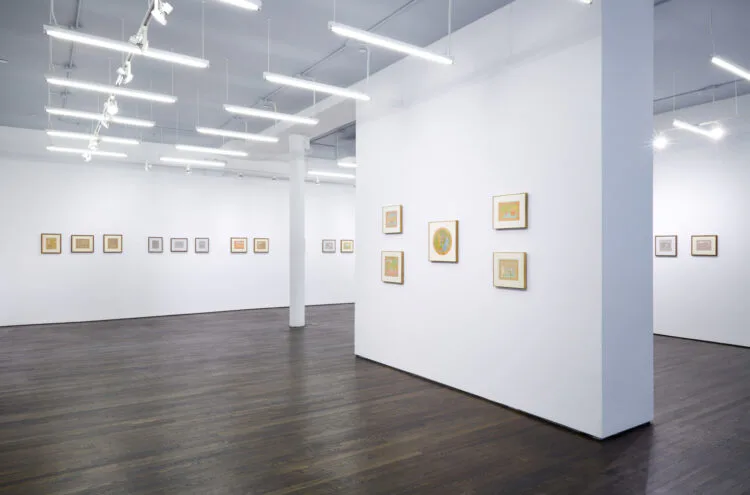 White gallery space with polished dark floor, support columns, and numerous small framed artworks arranged salon-style across multiple walls.