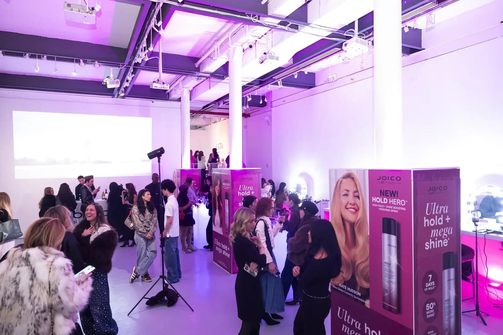 Gallery event with crowd under vibrant purple ambient lighting, branded displays, white columns, and projection screen in industrial venue.
