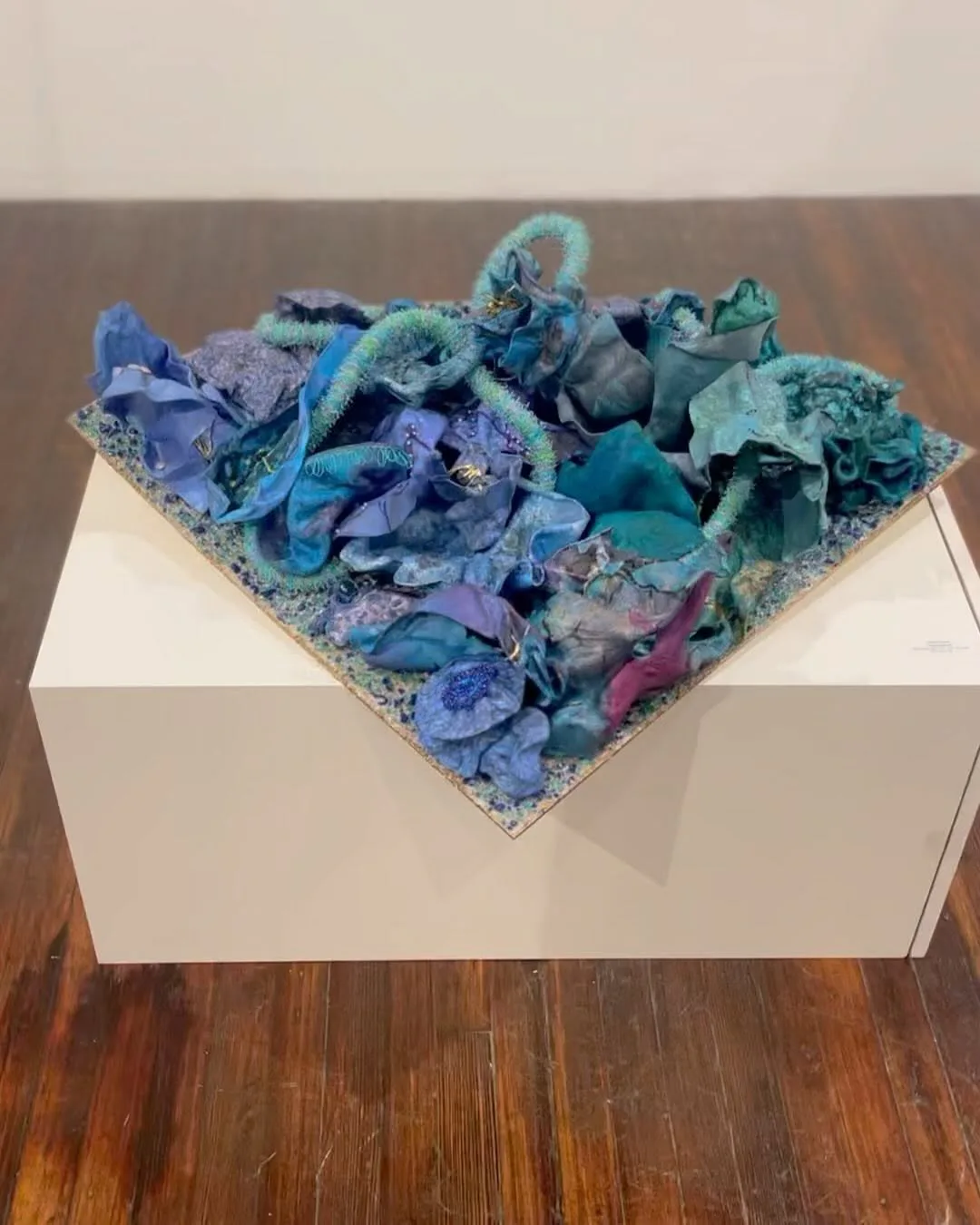 Three-dimensional mixed-media sculpture featuring layered turquoise, purple, and teal paper or fabric forms arranged on a white square base, displayed on wooden floor.