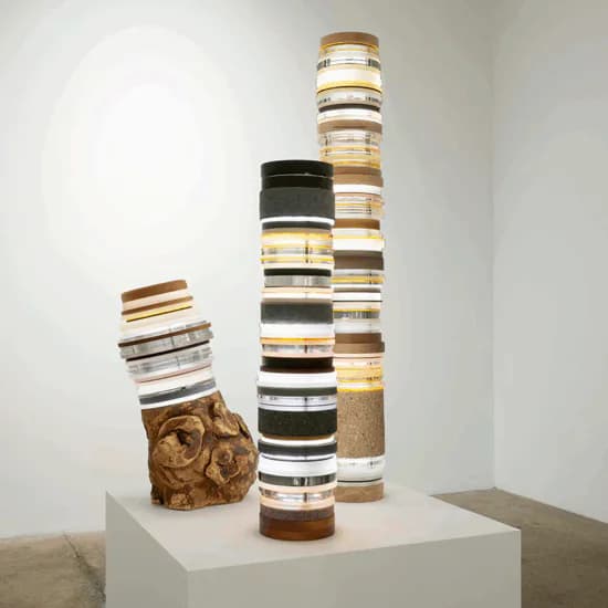 Sculptural installation on white pedestal featuring stacked cylindrical fabric rolls in cream, brown, and black stripes with a weathered golden crumpled form at the base.