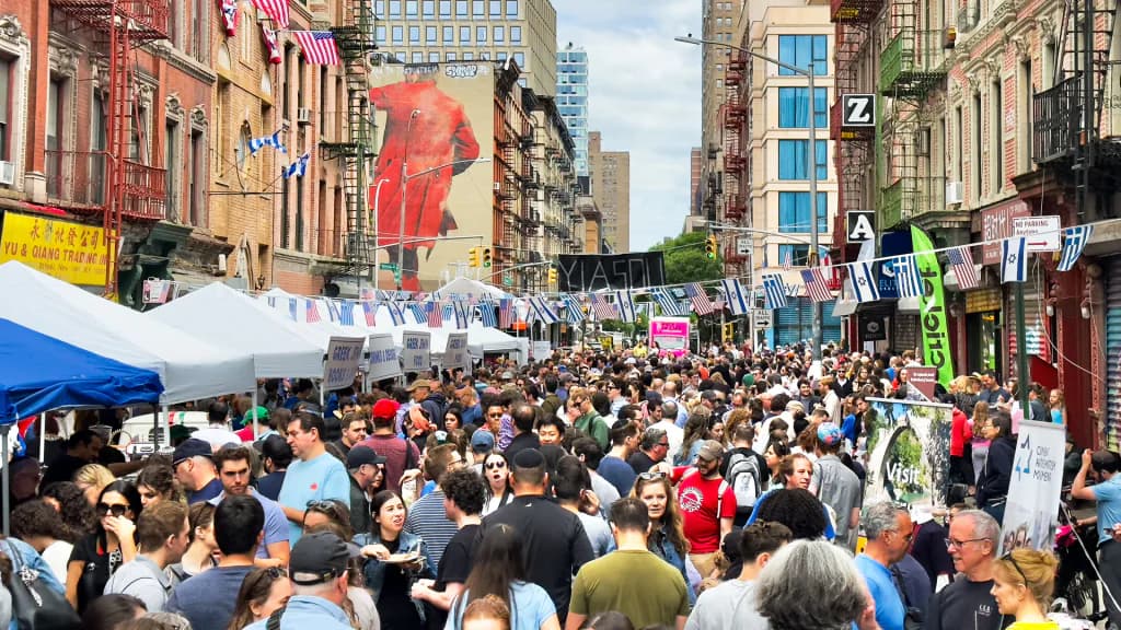 Crowded urban street festival scene with large red figure artwork on brick building, vendor tents, and numerous people gathered in lower Manhattan.