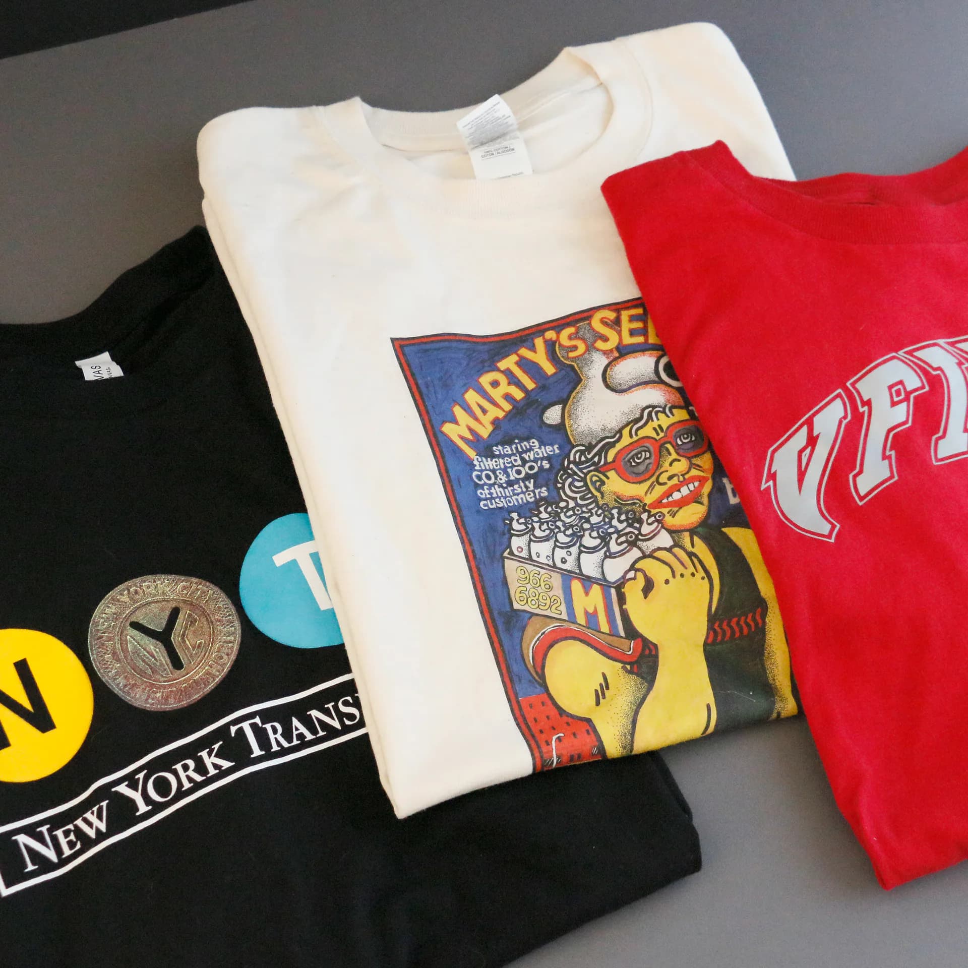 Collection of folded t-shirts displaying various graphic designs including vintage band imagery, cityscape graphics, and typography on cream, red, and black cotton.