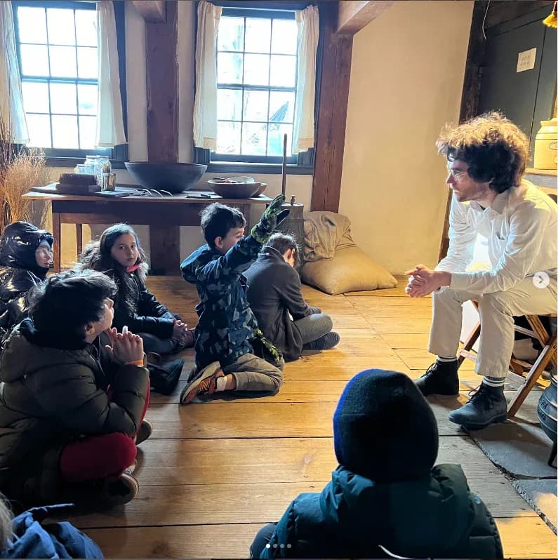 Children sit on a wooden floor in a bright room with large windows and exposed beams at Wyckoff House, listening to an adult educator in a cream-colored jacket.